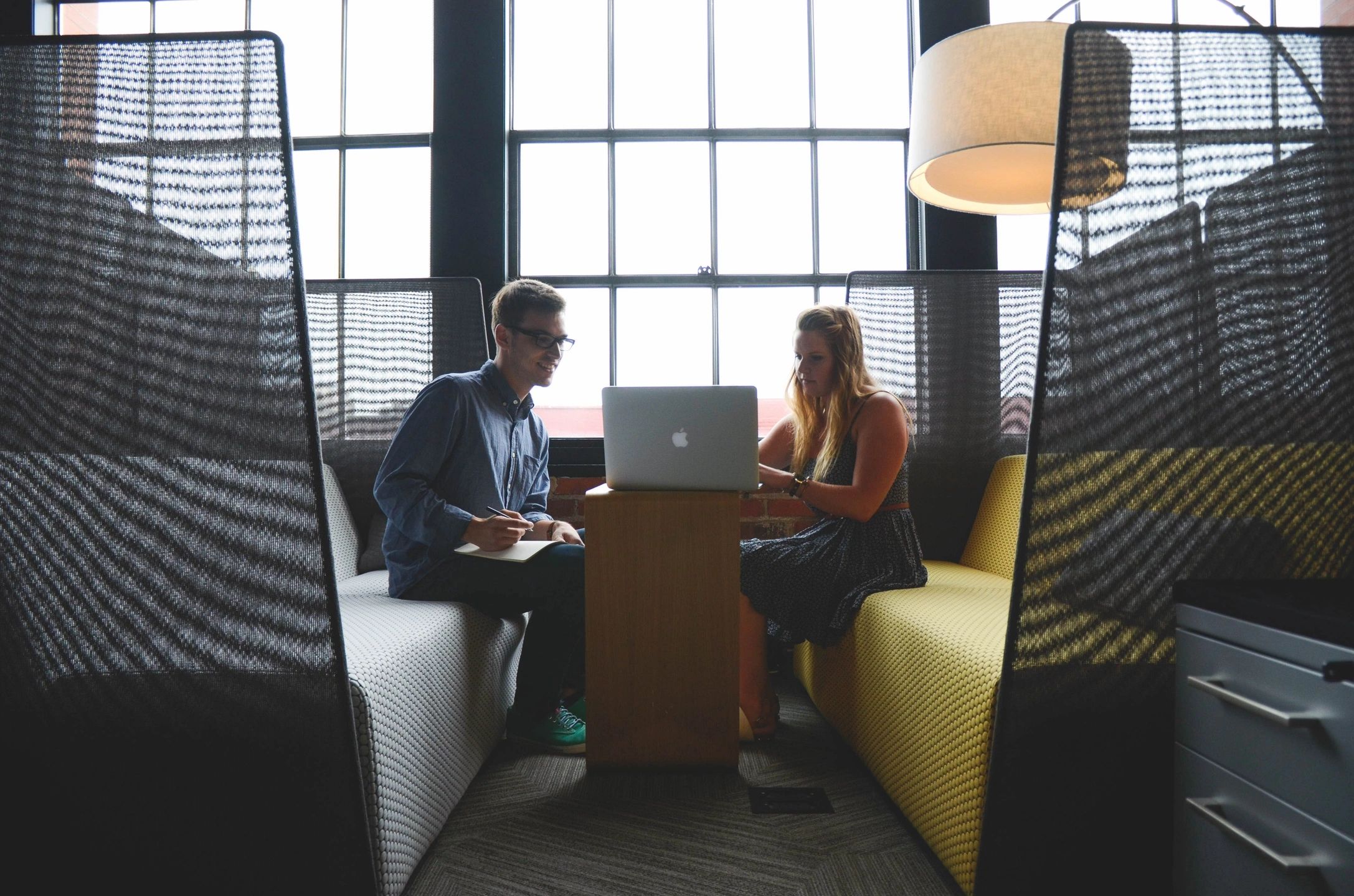 Two poeple sat opposite eachother in a modern work space, they have a laptop open that are are both looking at on a stoll in the middle of them